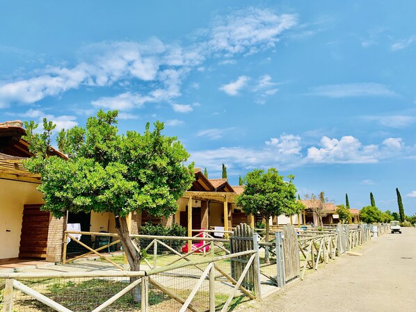 A row of traditional buildings with wooden roofs and a fenced area with trees. - Casa in Maremma Tuscany Village (Scarlino)