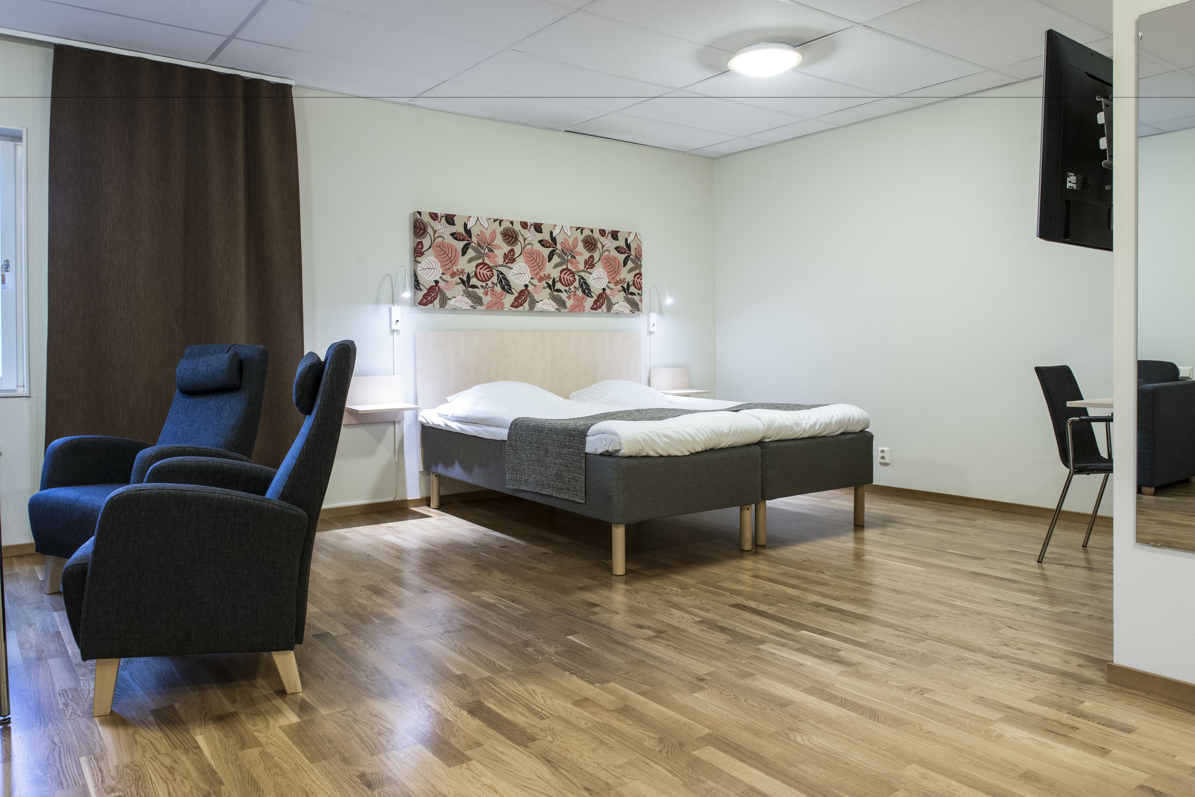 A modern hotel room with a bed, two armchairs, a desk, and a wall-mounted TV.