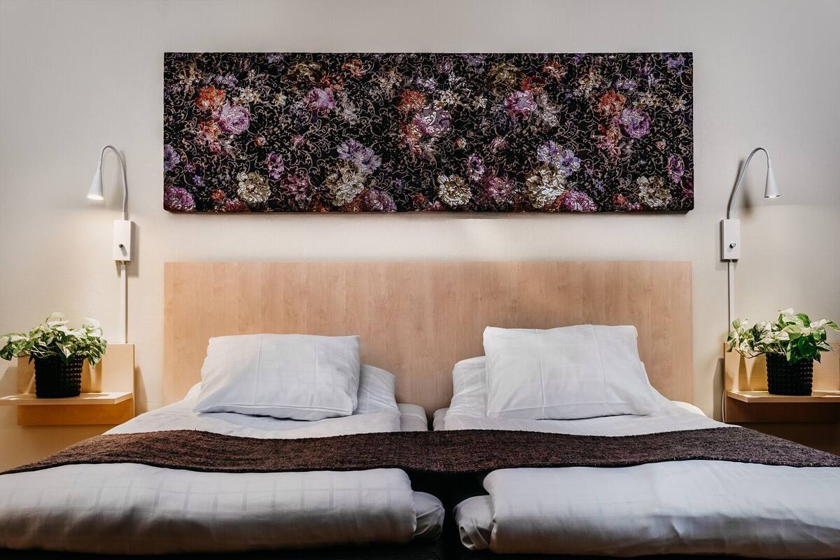 Habitación de hotel con cabecera de madera, dos almohadas blancas, una manta oscura y dos mesitas de noche con lámparas y plantas en macetas.