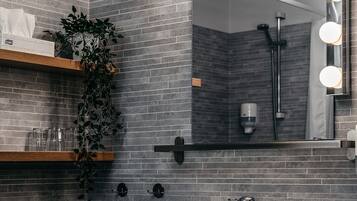 A bathroom with a grey tiled wall, a white sink, a mirror with lights, a plant, and a shelf with glasses.