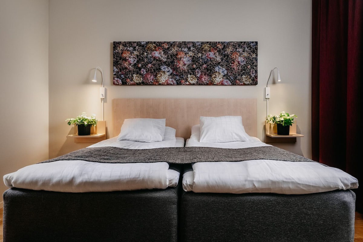 Una cama doble con ropa de cama blanca, dos mesitas de noche con lámparas, una obra de arte floral sobre la cama y una cortina roja a la derecha.