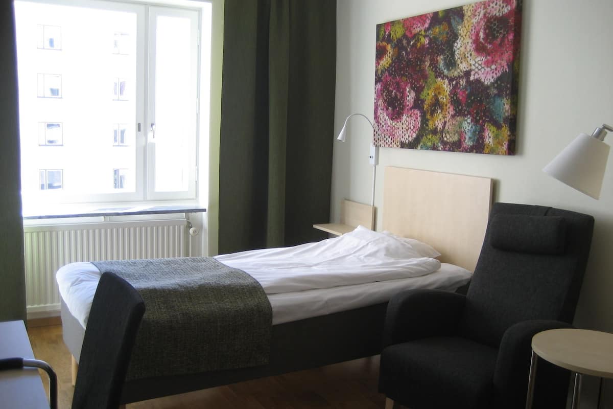 Habitación de hotel con cama, una silla, una mesita, una lámpara, un ventanal con cortinas y un cuadro abstracto de colores en la pared.