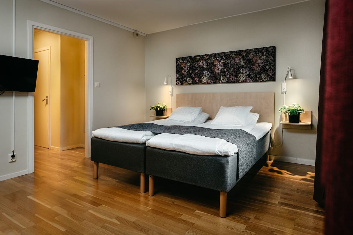 Una cama doble con cabecera oscura, dos mesitas de noche con lámparas, un televisor montado en la pared y suelo de madera.