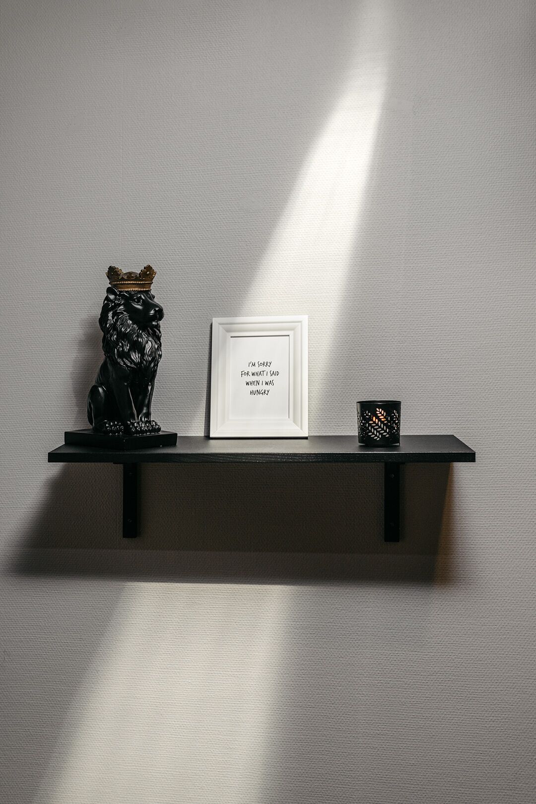 Estante de metal negro sobre una pared con textura, con una cita enmarcada, una figurita de león negro y un candelabro negro.