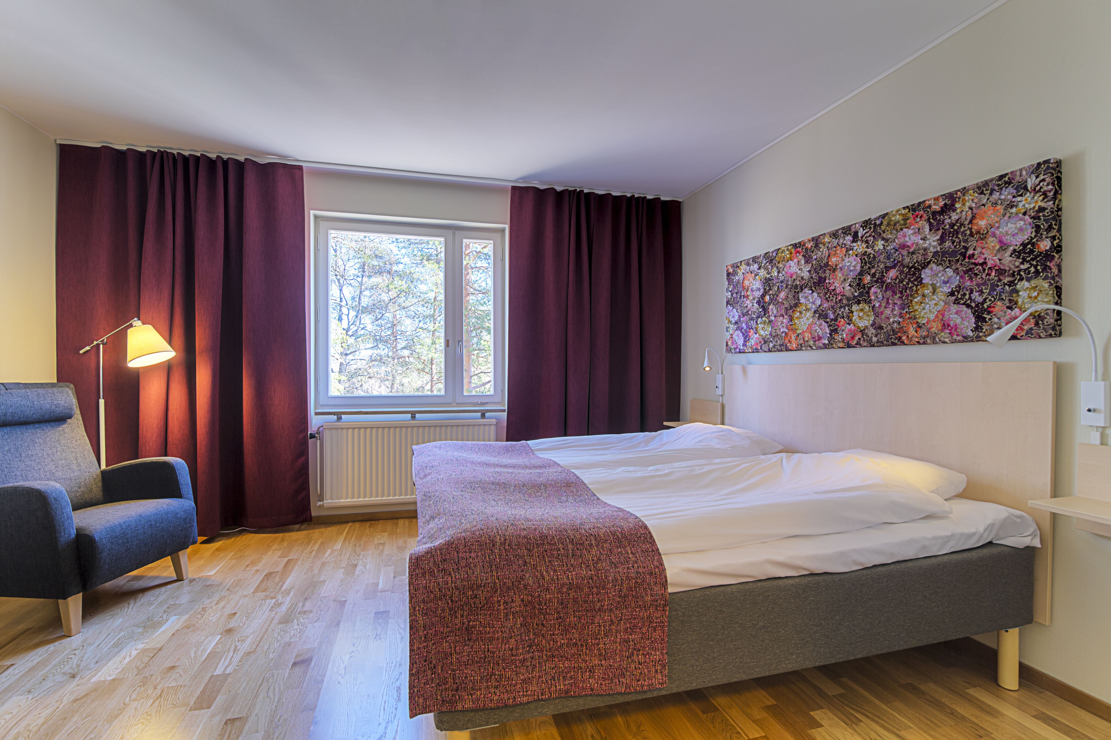 Habitación de hotel con cama, una silla, ventana con cortinas, radiador y un cuadro en la pared.