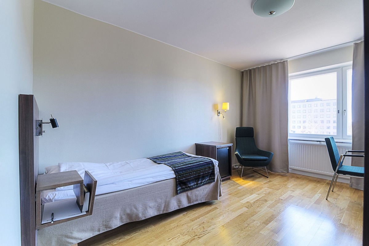 A hotel room with a bed, a desk, a chair, a lamp, and a window with curtains.