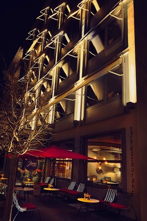 A building with multiple floors, outdoor seating with red umbrellas, and a visible restaurant sign. - Design-Hotel JAMS (Munich)