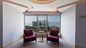 Habitación de hotel con dos sillas rojas, una mesita pequeña y vistas a una ciudad y montañas a través de ventanales amplios.