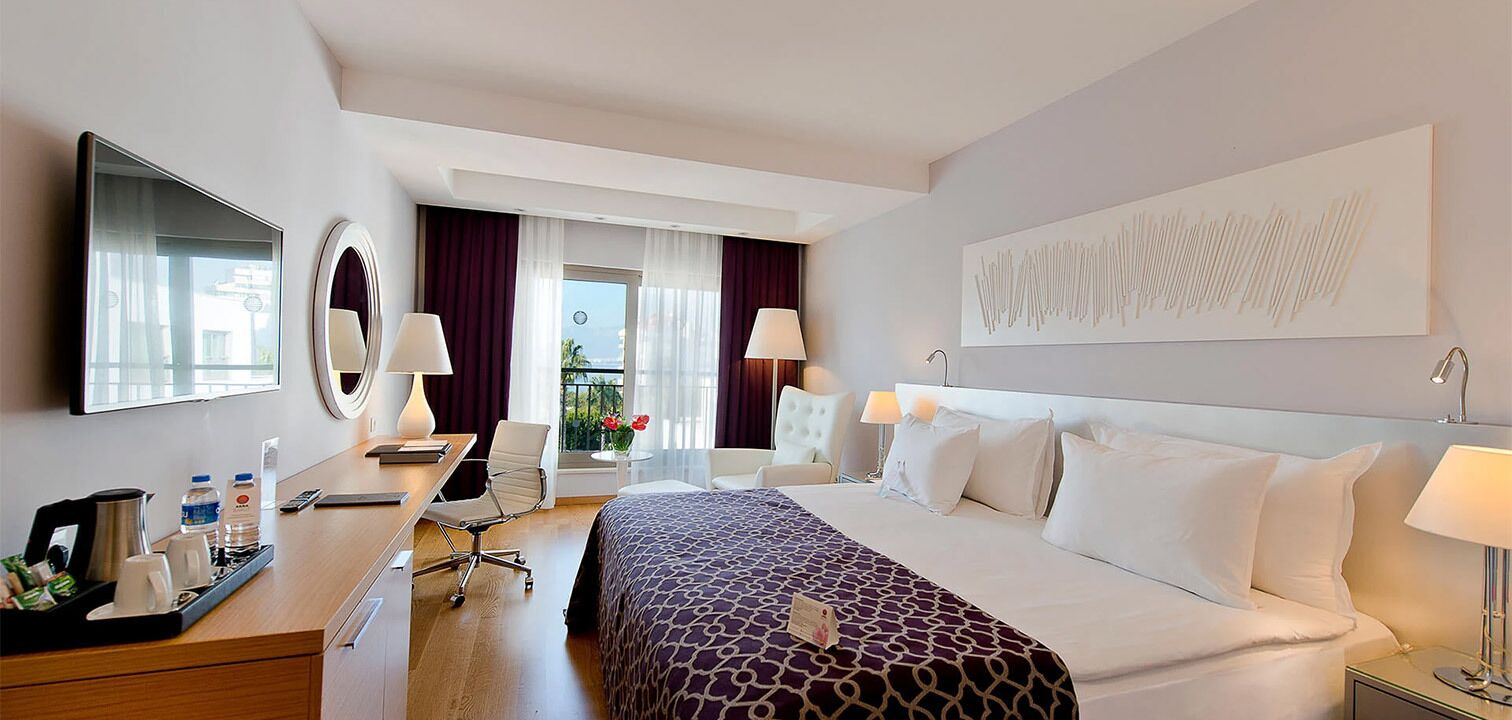 Habitación de hotel con cama, mesitas de noche, escritorio, silla, espejo, ventana con cortinas y una obra de arte colgada en la pared.