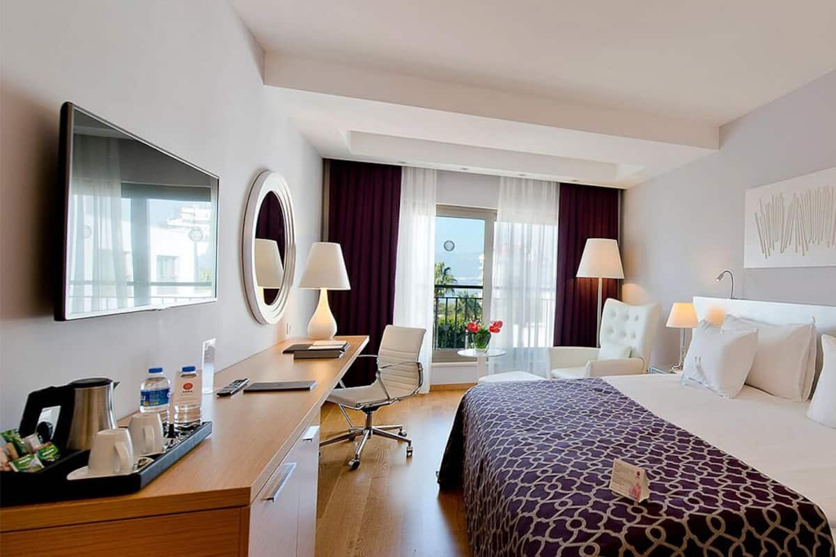 A hotel room with a bed, bedside tables, a desk, a chair, a mirror, a window with curtains, and a wall-mounted artwork.