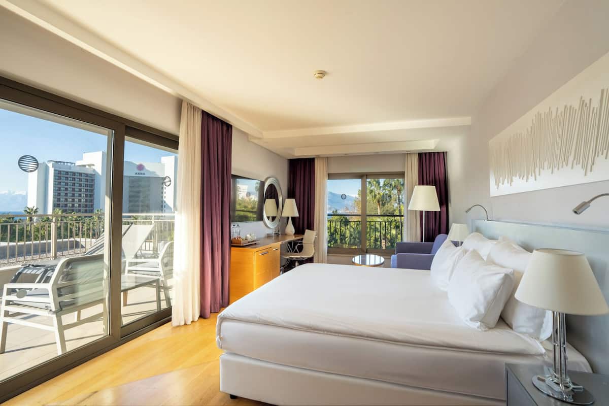 A hotel room with a large bed, a desk, a chair, a balcony with a view of the city and mountains, and a large window.
