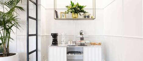 Une pièce moderne aux murs blancs, dotée d’un espace café, d’une plante en pot et d’un miroir au-dessus du comptoir.
