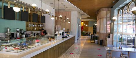 Una cafetería moderna con un mostrador de vidrio que exhibe pasteles, techo de madera y zona de estar con mesas y sillas.