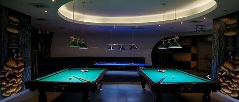 Espaço com mesas de bilhar, duas unidades, iluminação pendente e parede com padrão xadrez.