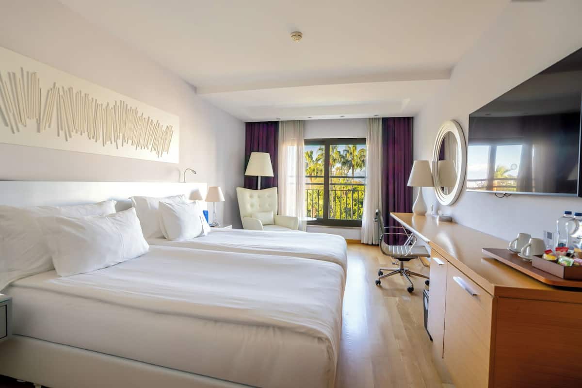 A hotel room with two beds, a desk with a mirror, a chair, and a view of palm trees through the window.