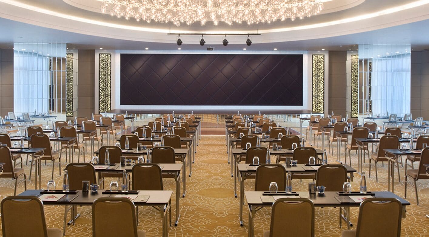 A large conference room with rows of tables and chairs, a large screen at the front, and a chandelier above.