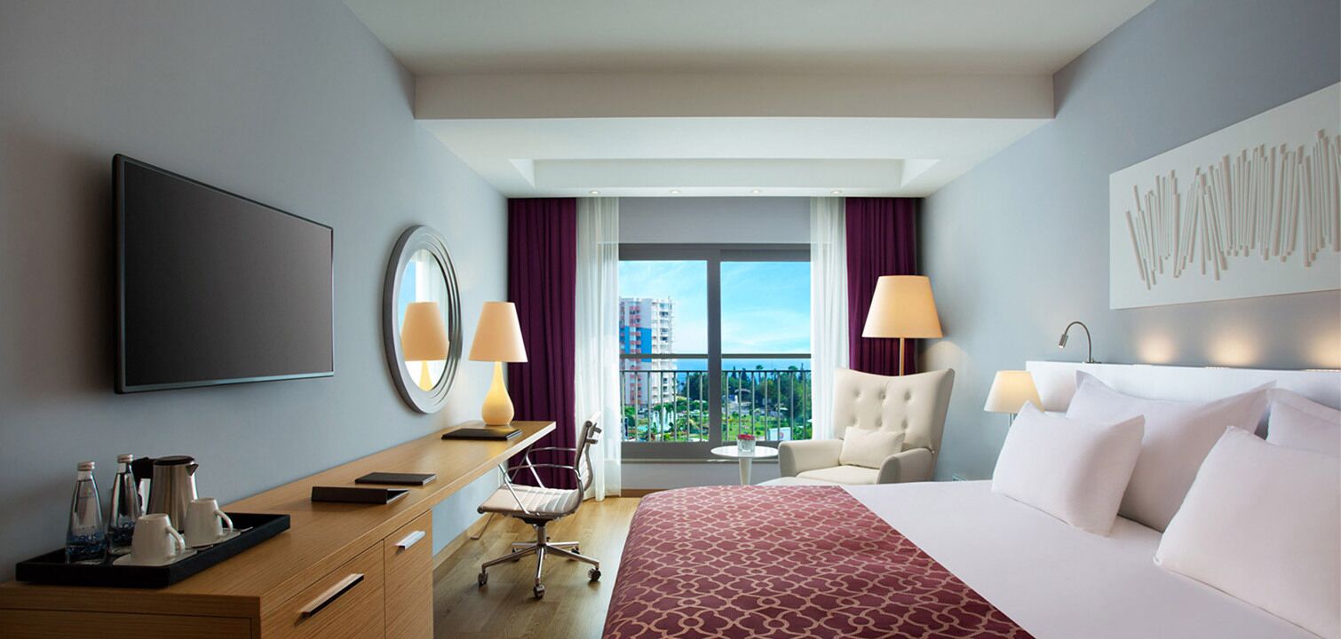 Habitación de hotel con cama, escritorio y silla, televisión, ventana con cortinas y vista a edificios.