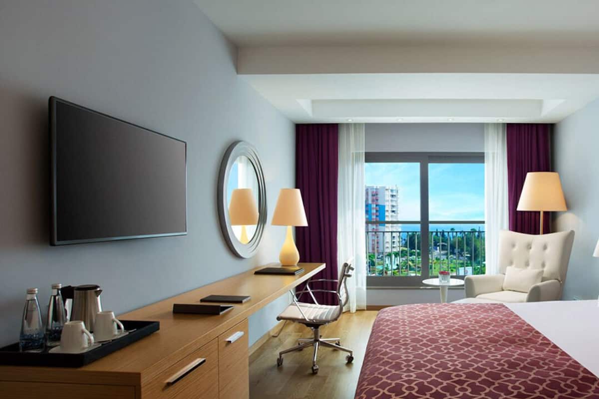 A hotel room with a bed, a desk with a chair, a TV, a window with curtains, and a view of buildings.