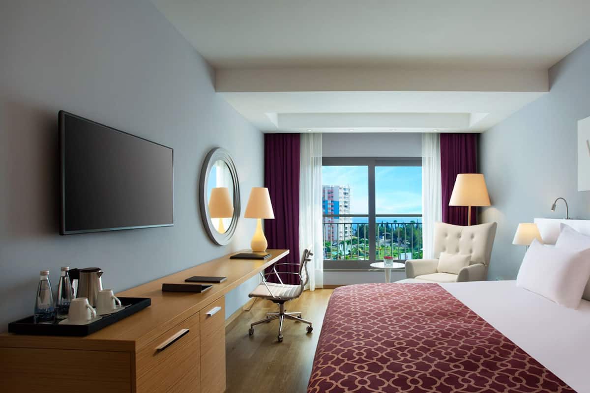 A hotel room with a bed, a desk with a flat-screen TV, a chair, a small table, and a view of the city.