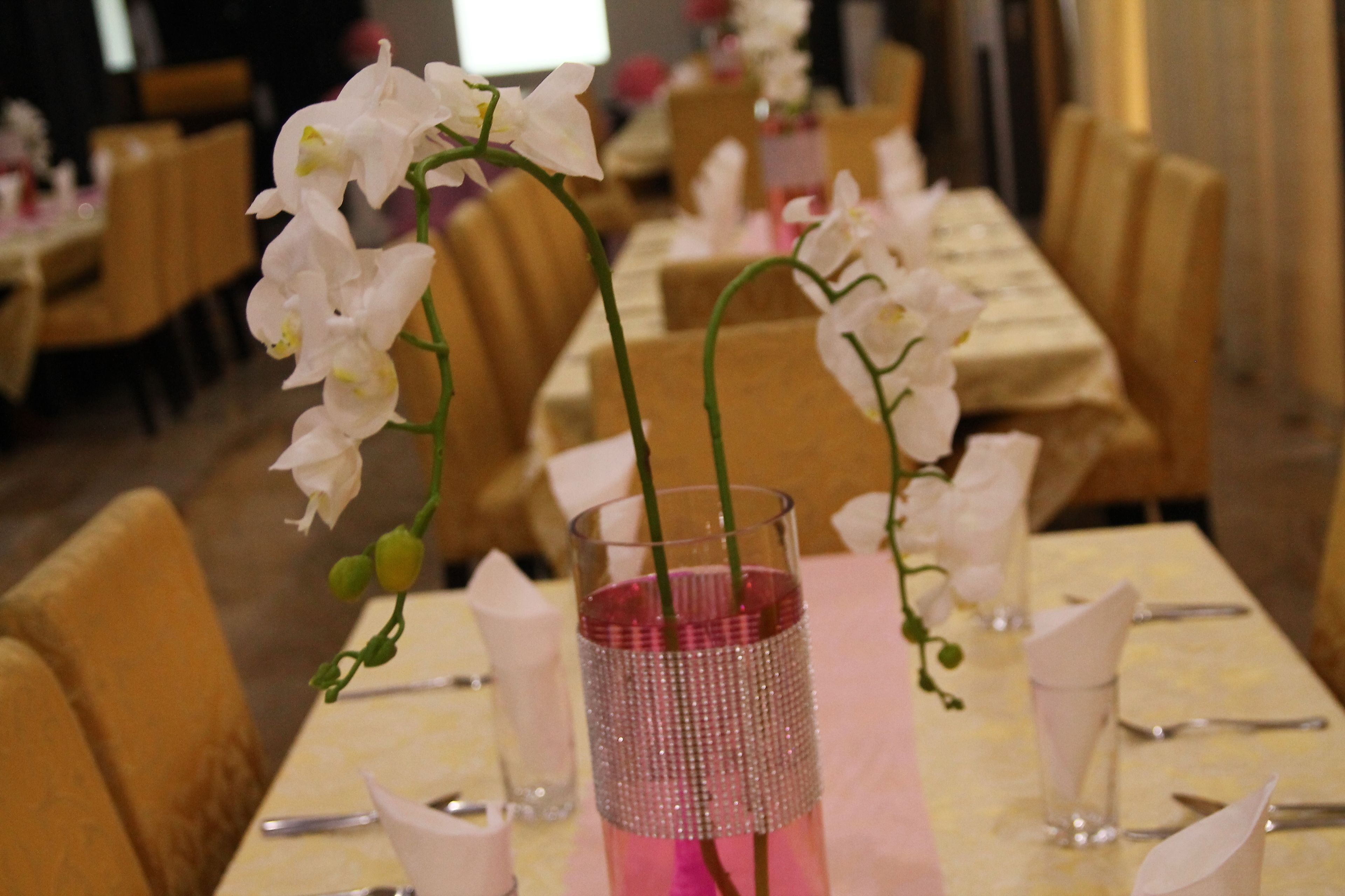 Uma mesa de jantar com um vaso rosa e orquídeas brancas, rodeada por cadeiras e louças bem organizadas.