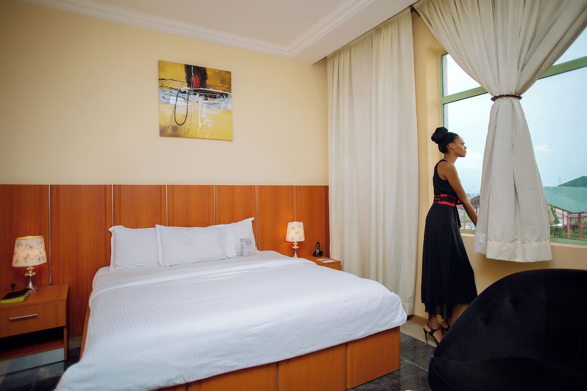 Een hotelkamer met een groot bed, een nachtkastje met lamp, een schilderij aan de muur en uitzicht door het raam.