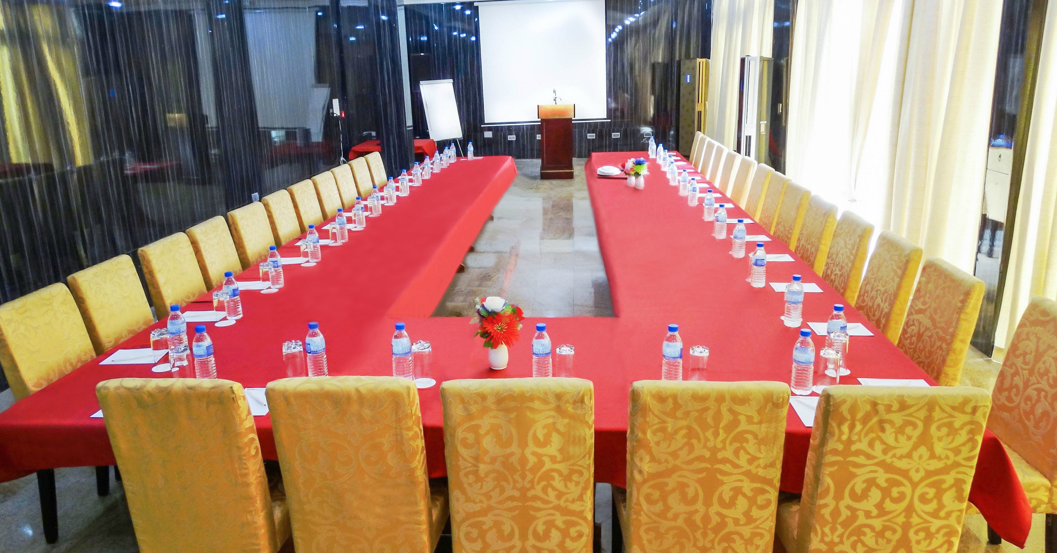 Uma sala de conferências com uma mesa comprida, cadeiras amarelas, garrafas de água e uma tela de projetor.