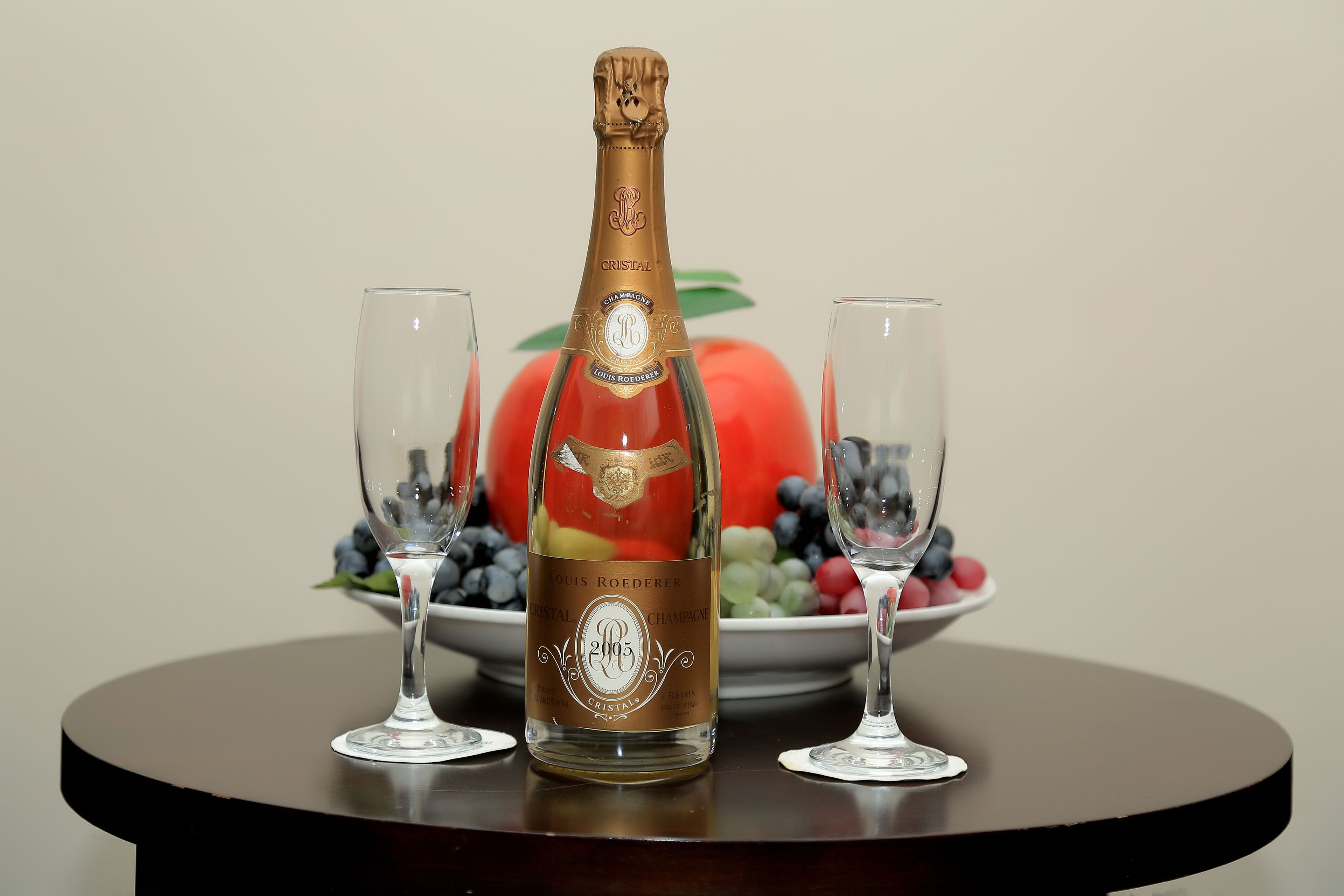 A bottle of Cristal champagne with two glasses on a table, with a bowl of fruit in the background.