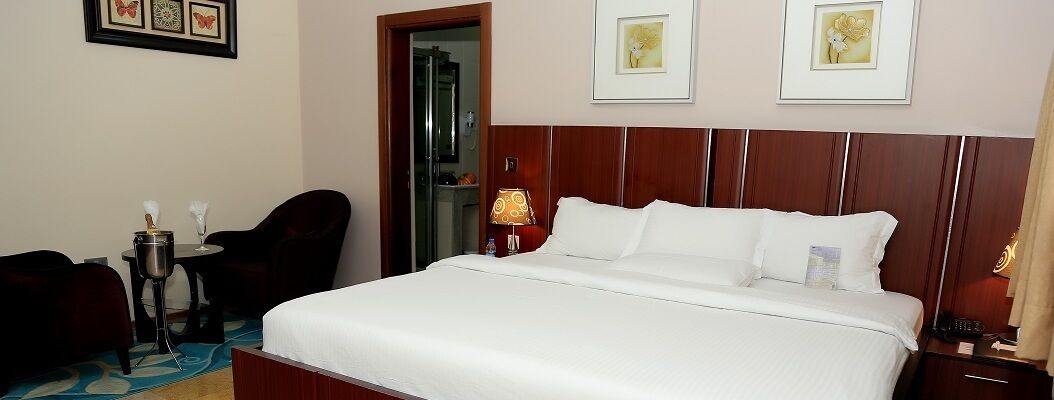 Habitación de hotel con una cama grande, un escritorio con silla, una mesita de noche con lámpara y cuadros en la pared.