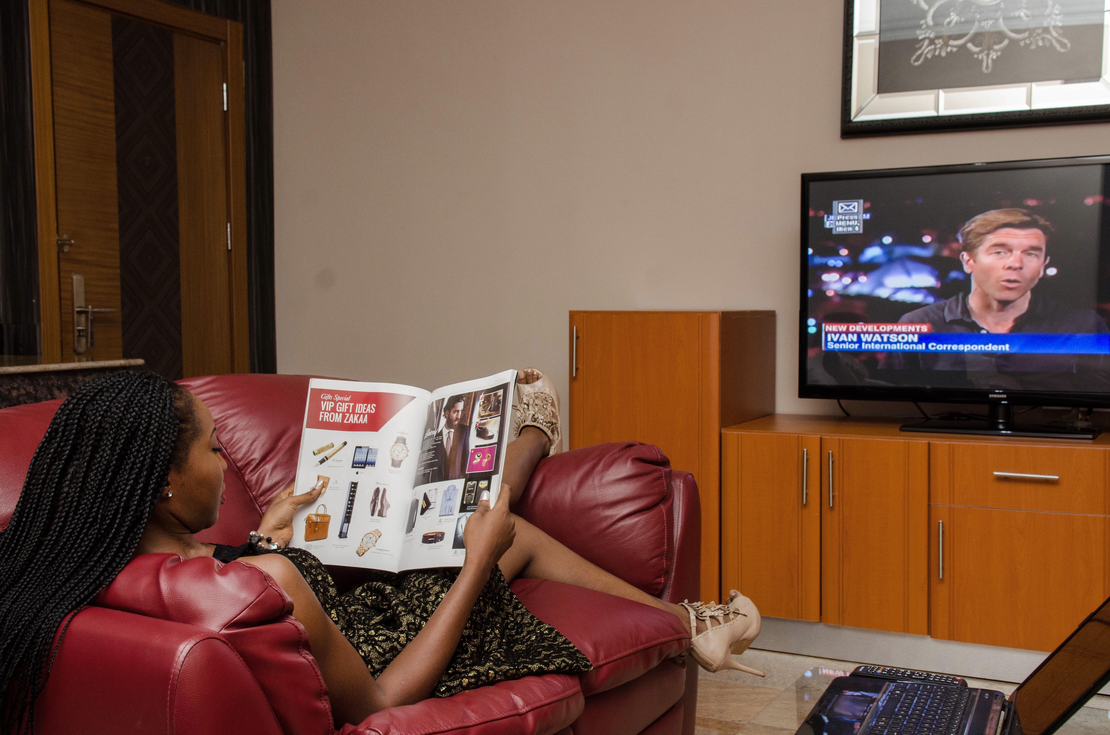 A person sitting on a red leather couch reading a magazine. A television is on a wooden stand to the right, displaying a news program.