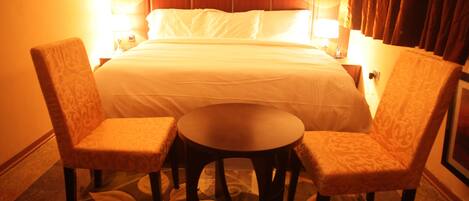 Habitación de hotel con una cama, dos sillones, una mesita redonda y una mesita de noche con una lámpara.