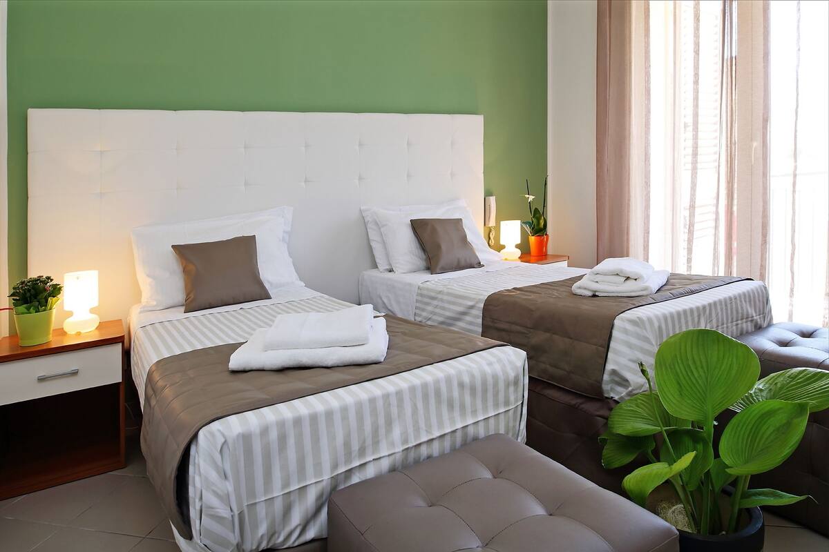 Camera d'albergo con due letti, un comodino con lampada, una pianta in vaso e una finestra con tende.