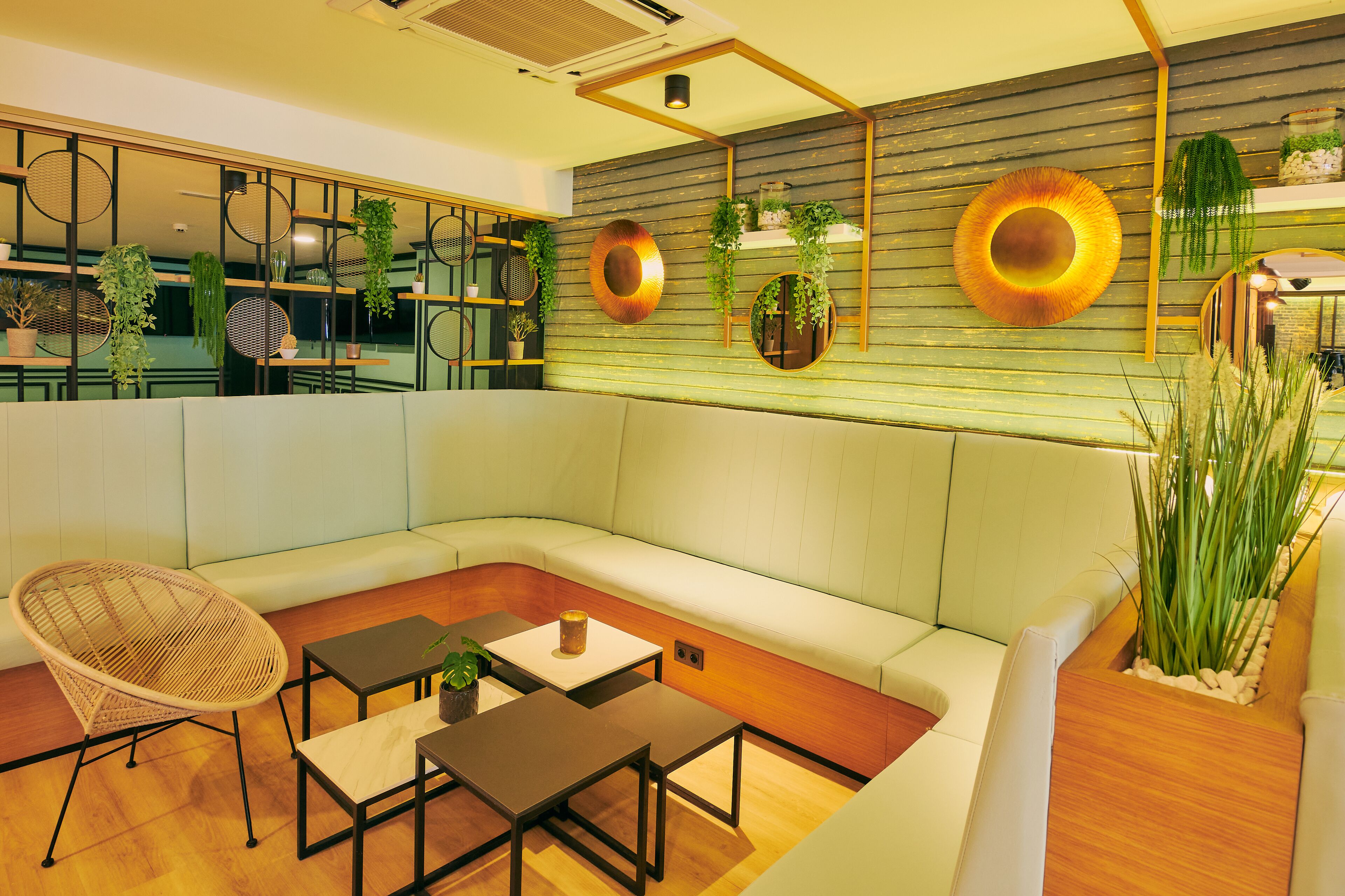 A modern lounge area with a sofa, two tables, a wicker chair, and decorative wall elements including plants and circular mirrors.