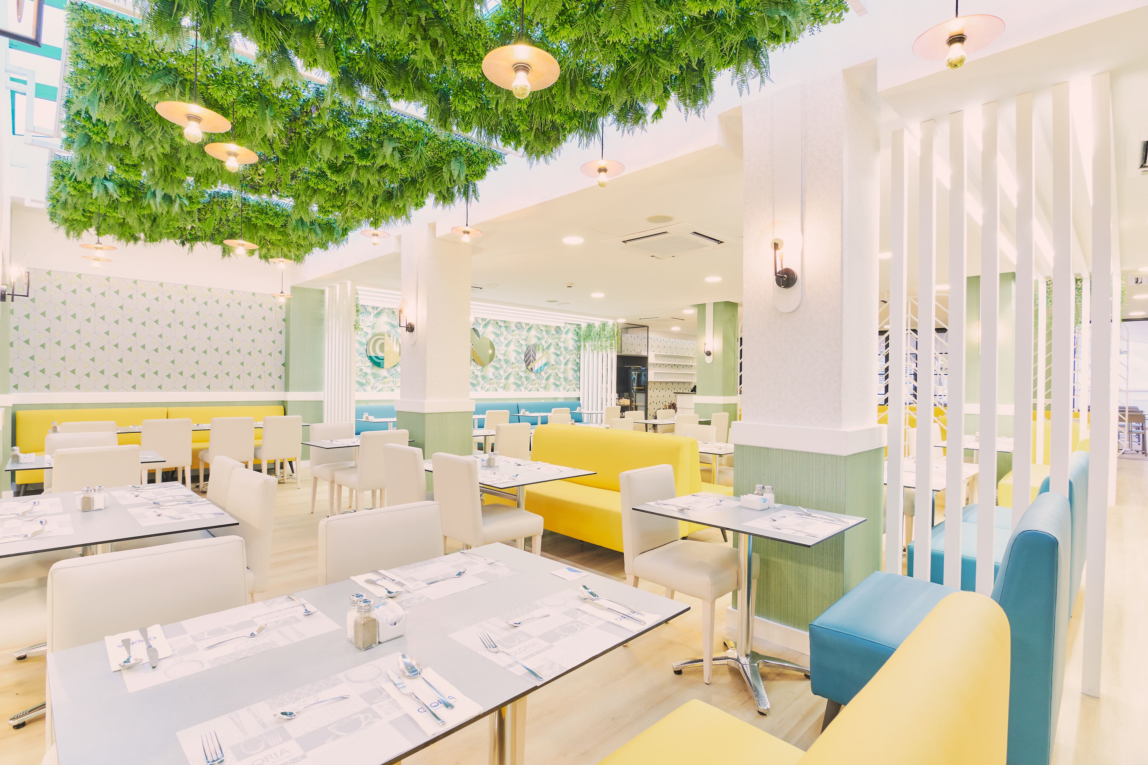 A restaurant with white tables, yellow and blue seating, and green hanging foliage.