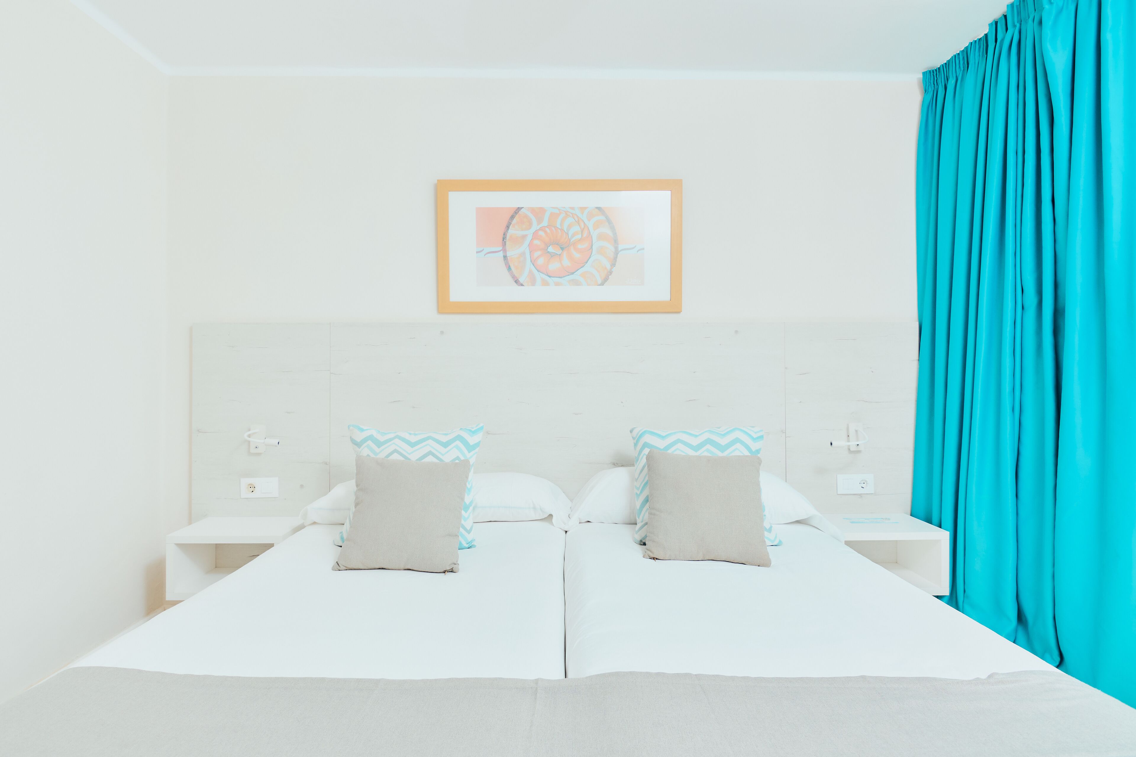 A double bed with two pillows, a nightstand, a framed picture on the wall, and a teal curtain.
