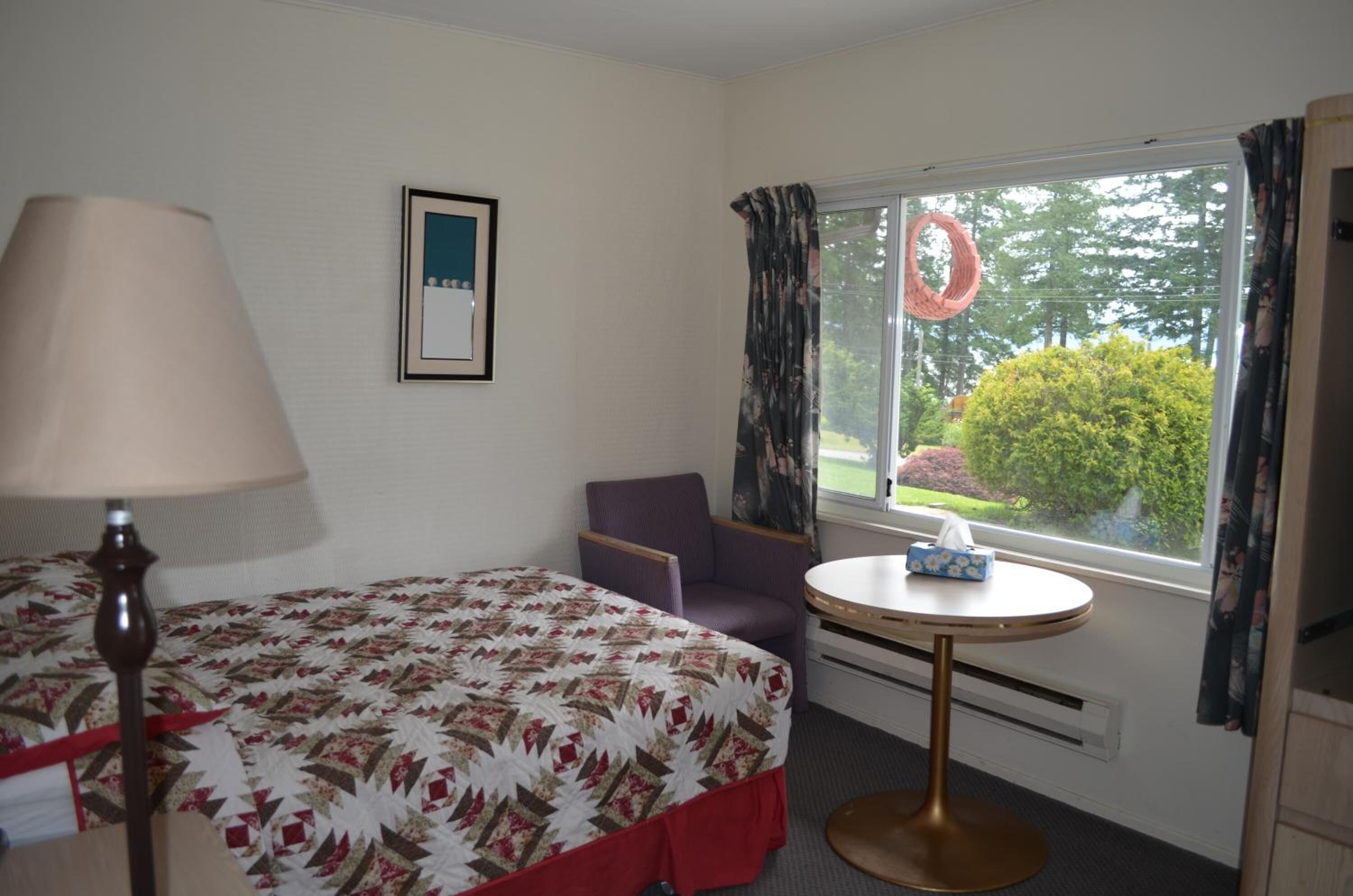 A hotel room with a bed, a chair, a small table with a tissue box, a lamp, and a window with curtains.