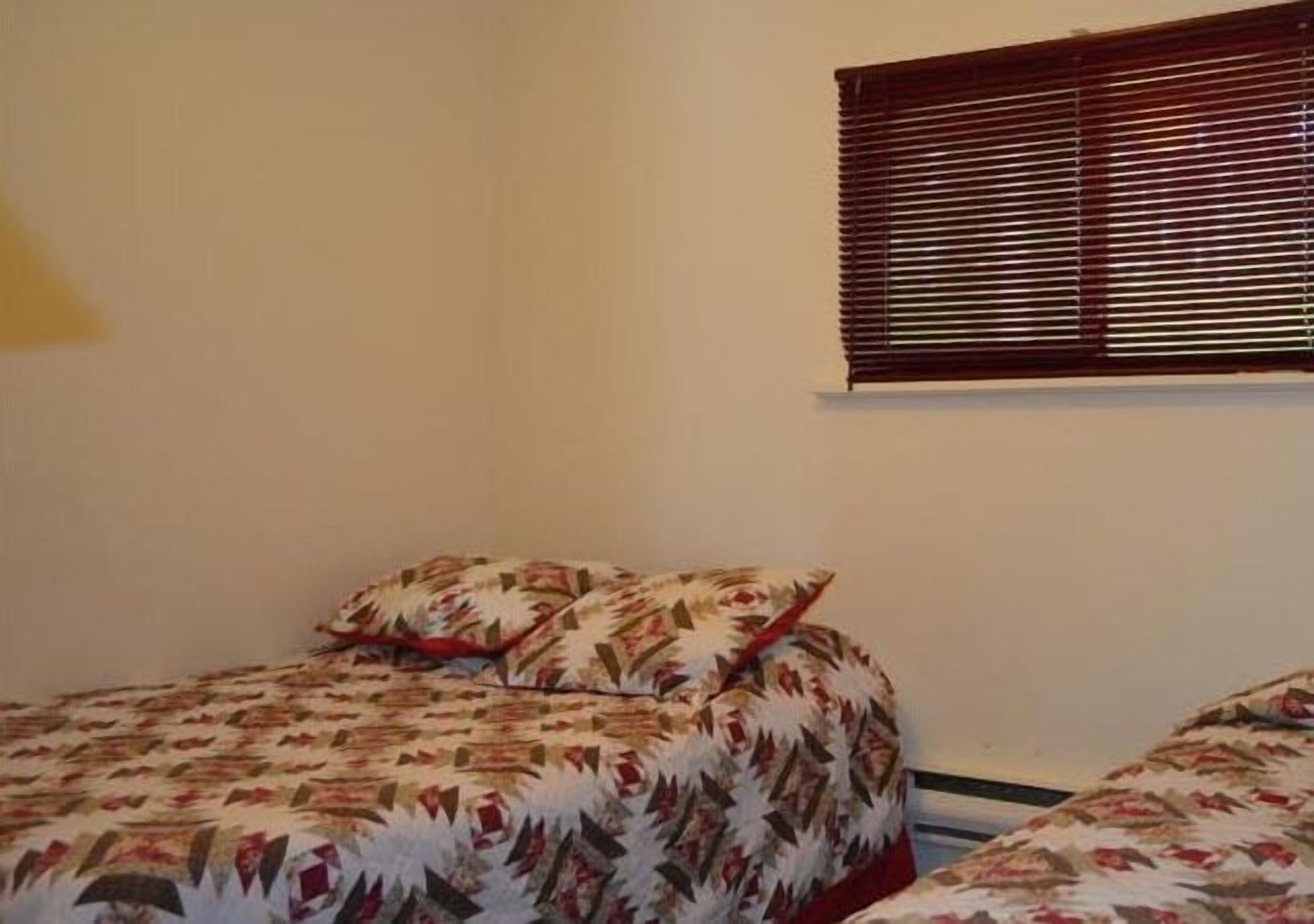 A bedroom with two beds, each with patterned bedding. There is a window with blinds on the wall.