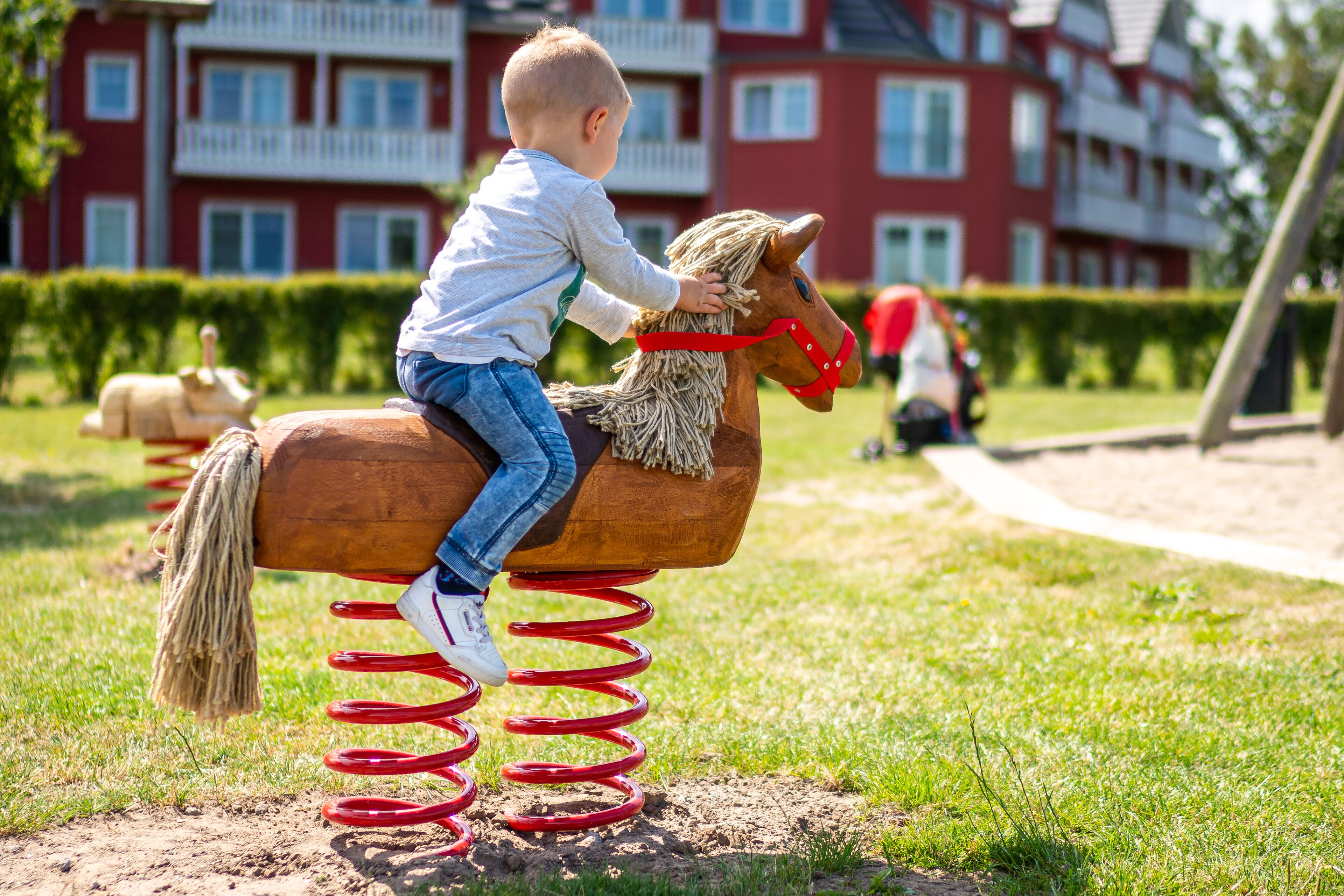 A child on a wooden horse with a red spring mechanism, set against a backdrop of a red building with balconies.