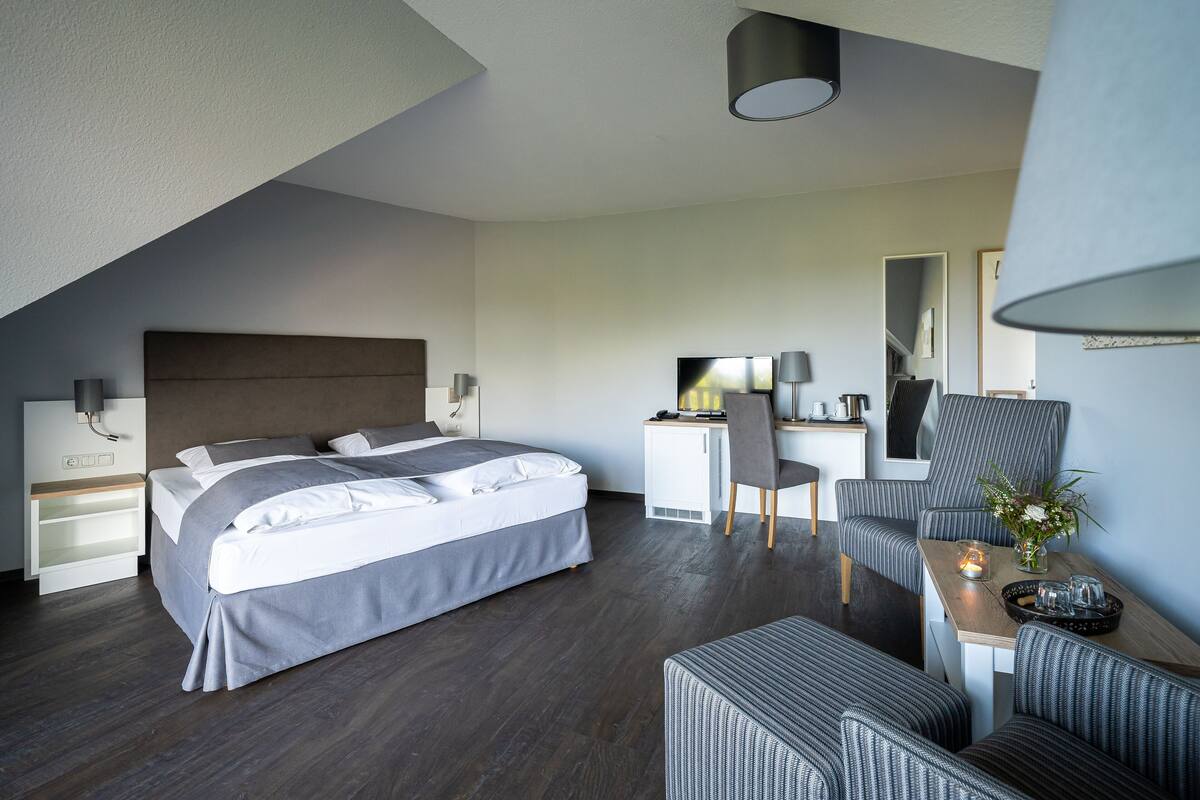 A modern hotel room with a large bed, a desk, a chair, and a dining area.