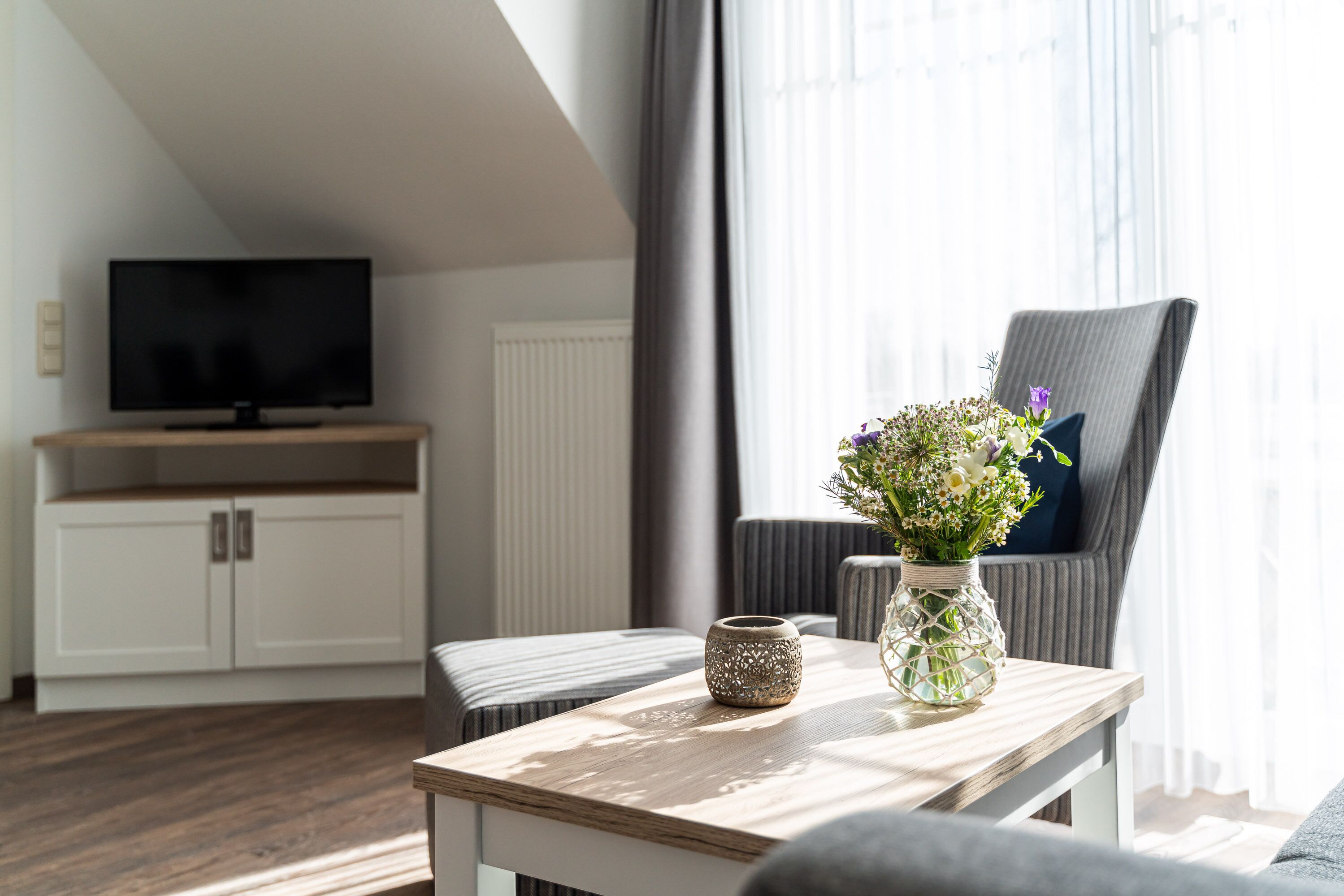 Una sala de estar moderna con un televisor de pantalla plana, una mesa de centro de madera, un jarrón decorativo con flores, un sillón gris y un armario blanco.