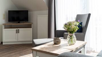 Una sala de estar moderna con un televisor de pantalla plana, una mesa de centro de madera, un jarrón decorativo con flores, un sillón gris y un armario blanco.