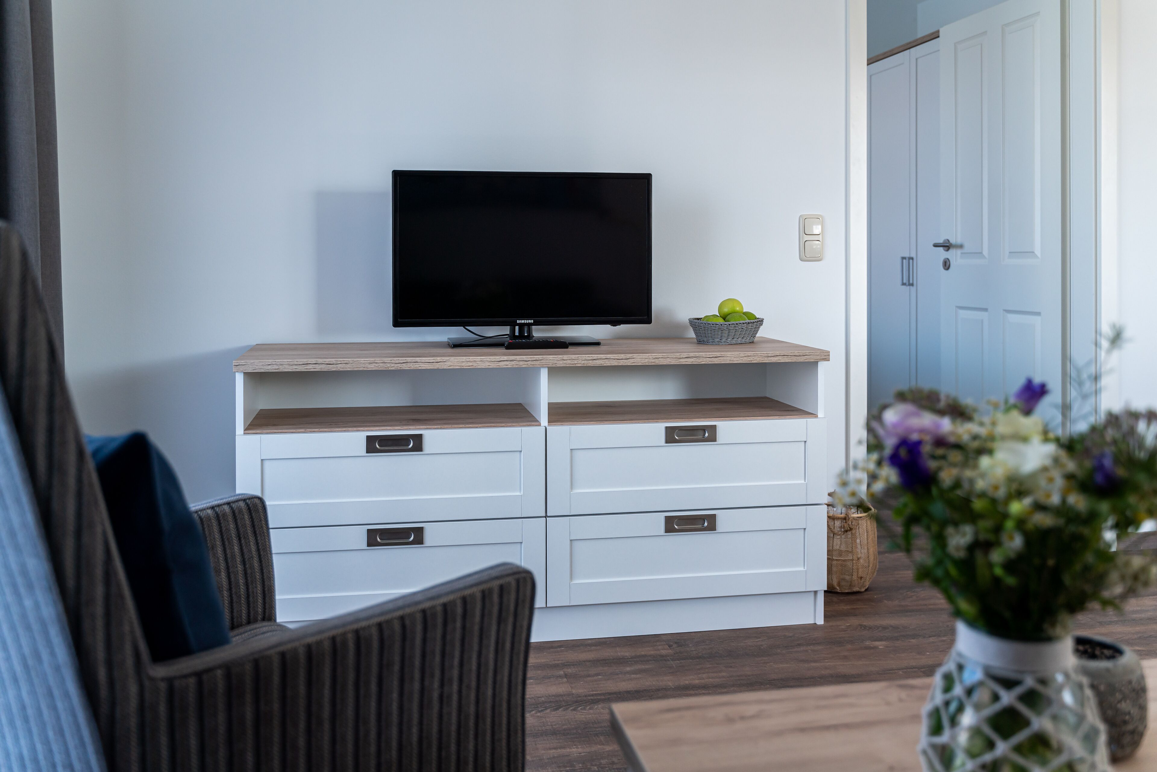 A modern living room with a flat-screen TV on a wooden shelf, white storage units, a wicker basket, and a vase with flowers.