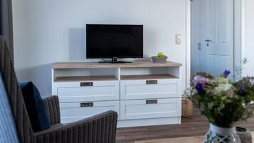 A modern living room with a flat-screen TV on a wooden shelf, white storage units, a wicker basket, and a vase with flowers.