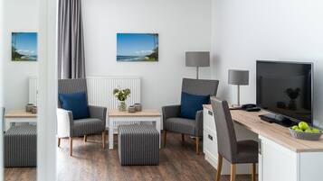 A modern hotel room with a wooden floor, two grey upholstered chairs, a small table with a vase, a desk with a computer, a lamp, and a TV.