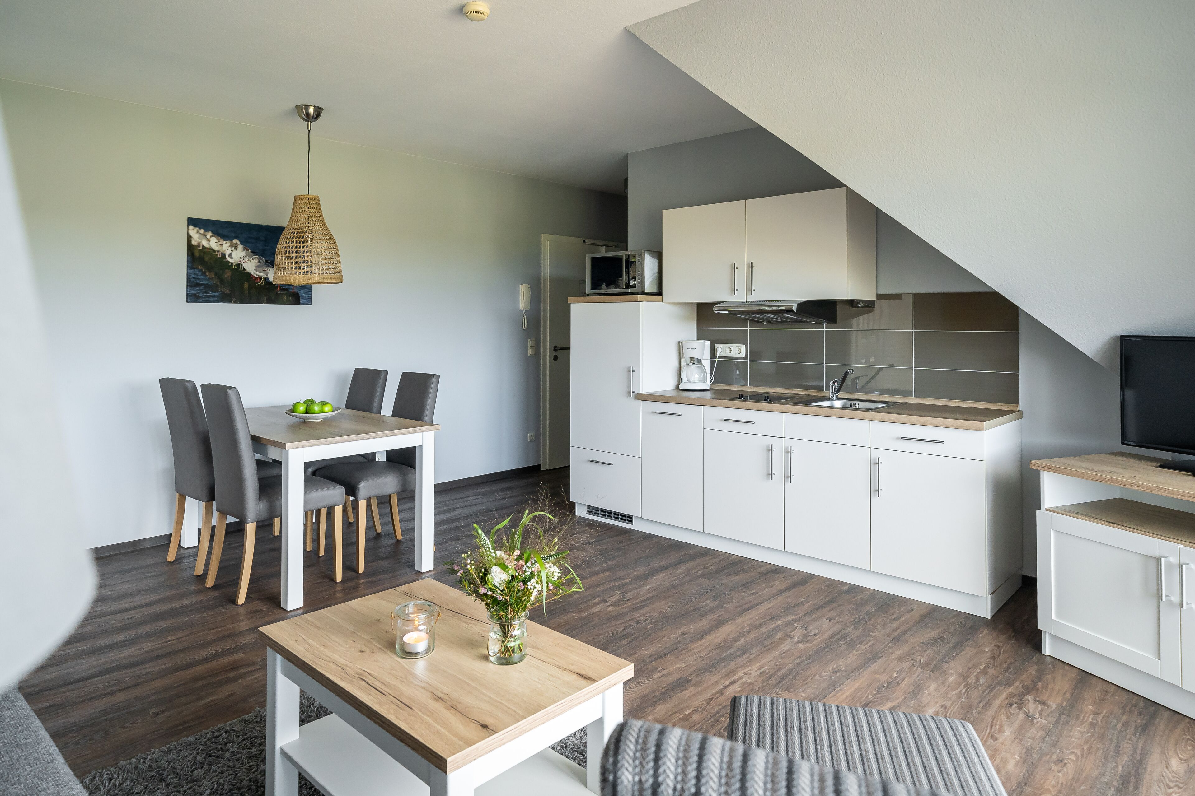 Een moderne keuken met witte kasten, een magnetron en een koffiezetapparaat. Er is een eethoek met een houten tafel en stoelen.