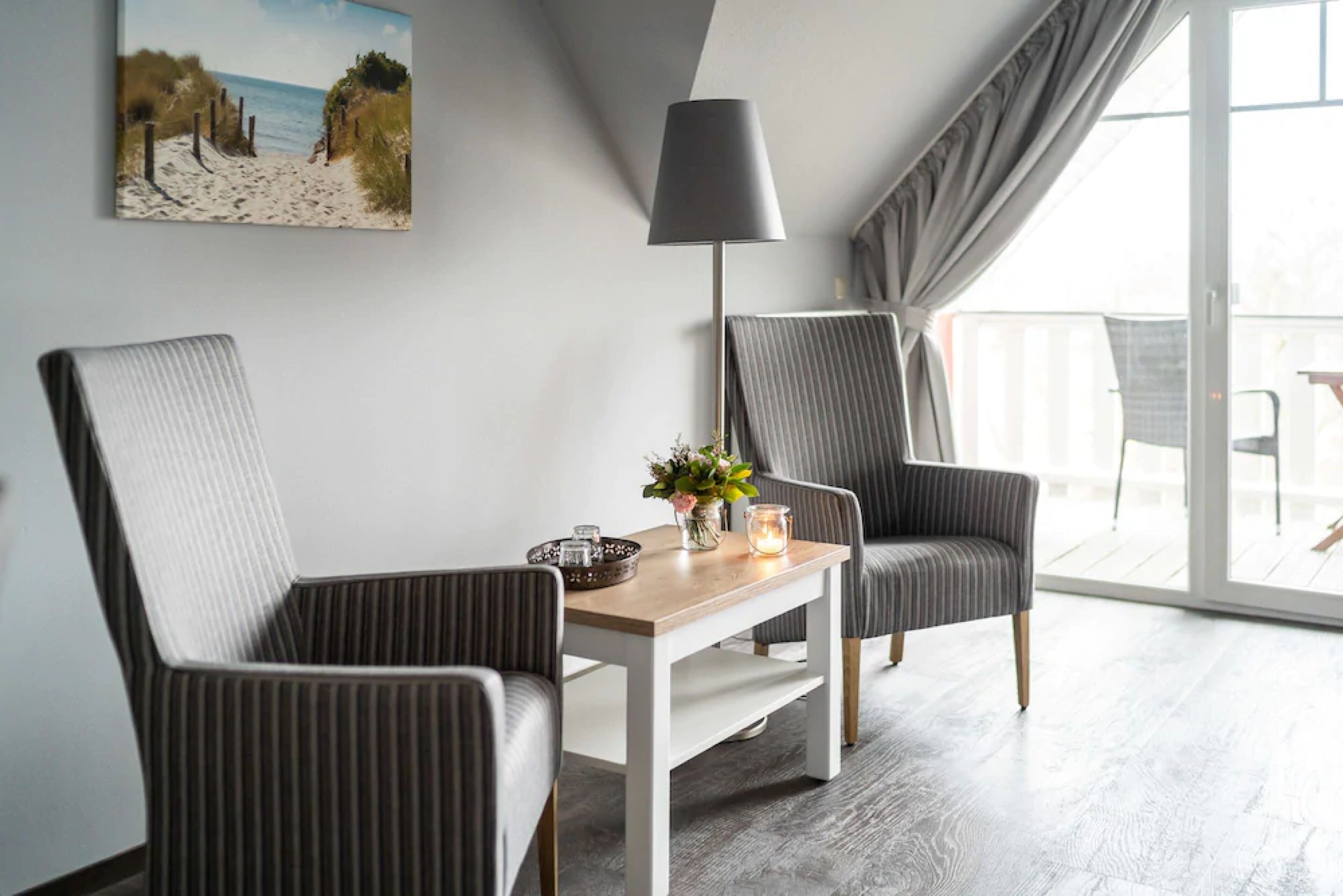A modern room with two upholstered chairs, a wooden table, a floor lamp, and a beach-themed painting on the wall.