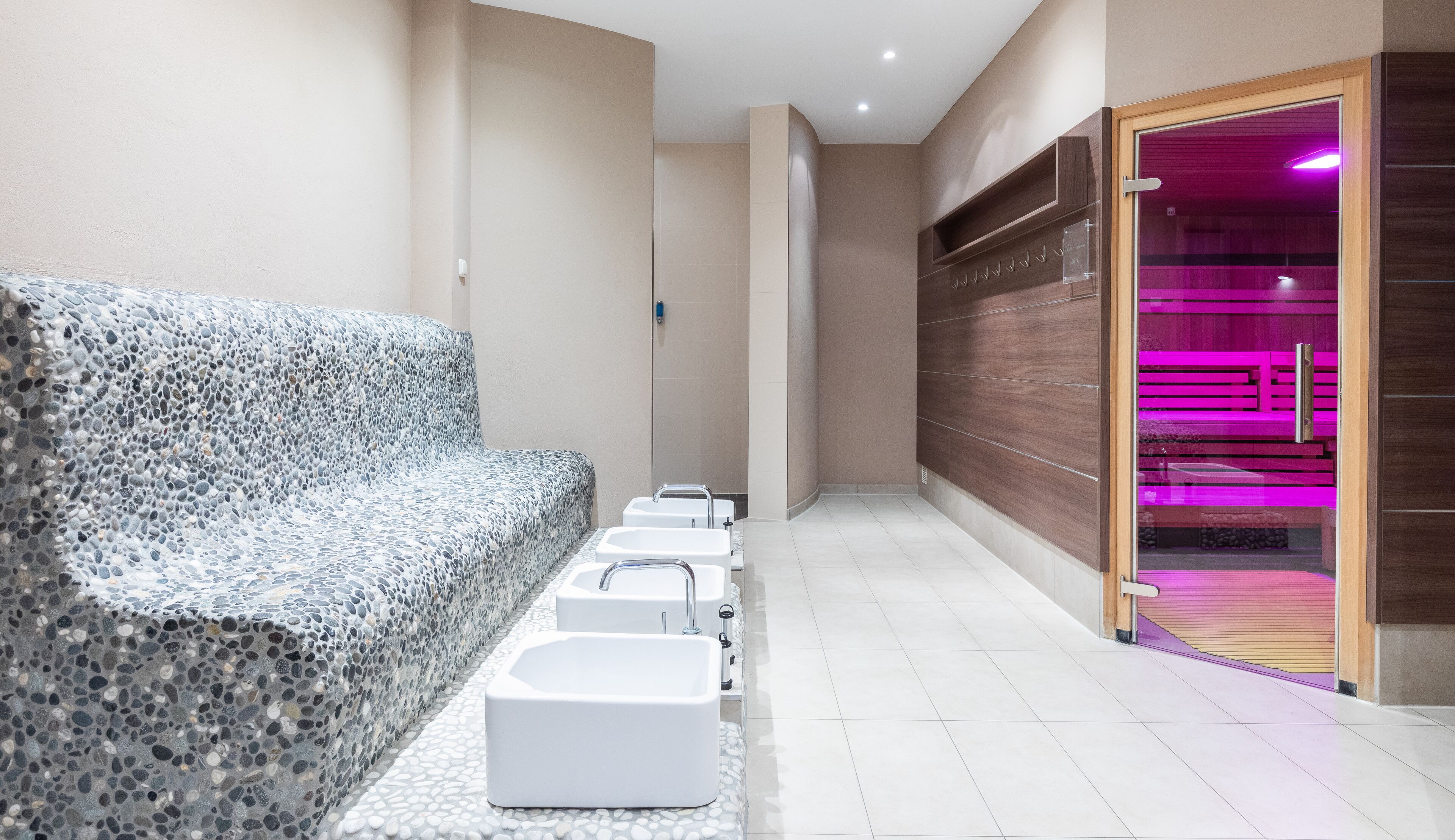 Uma área de spa moderna com parede texturizada em pedra, uma fileira de pias brancas e uma porta de madeira que dá para um espaço com iluminação rosa.