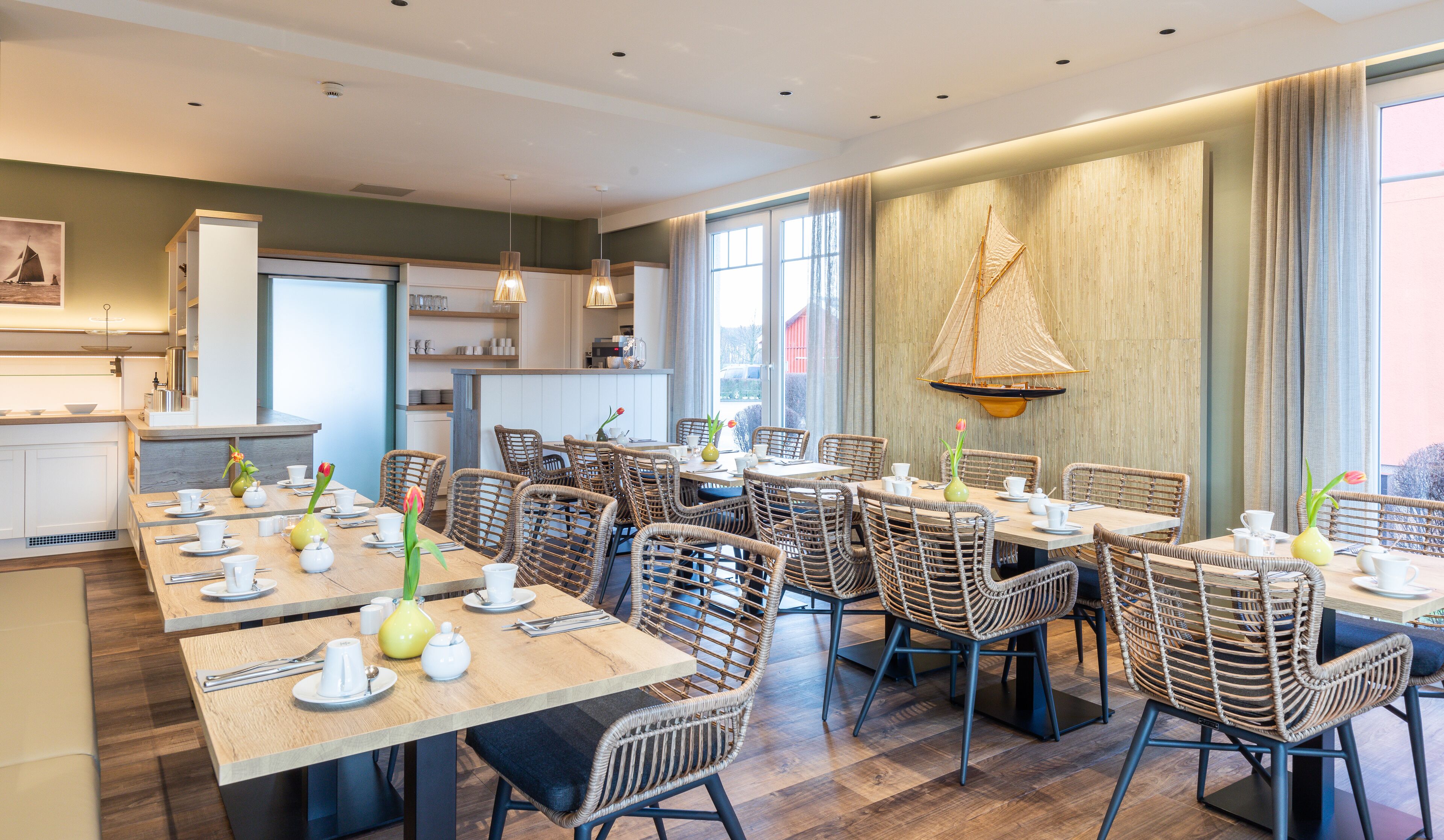 A dining area with multiple tables and wicker chairs, a kitchen in the background, and a large window with curtains.
