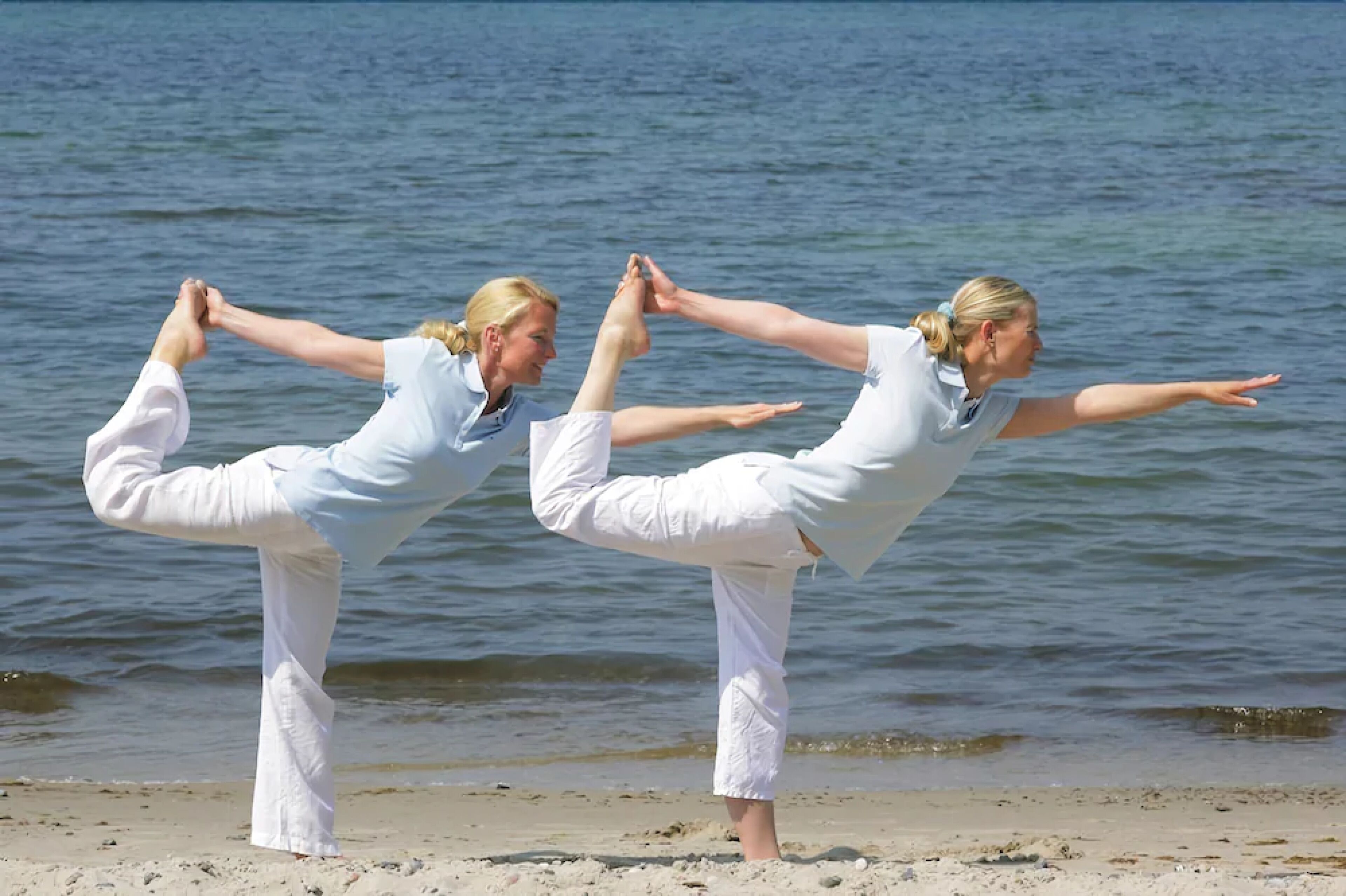 Two individuals performing a yoga pose on a beach with the ocean in the background.