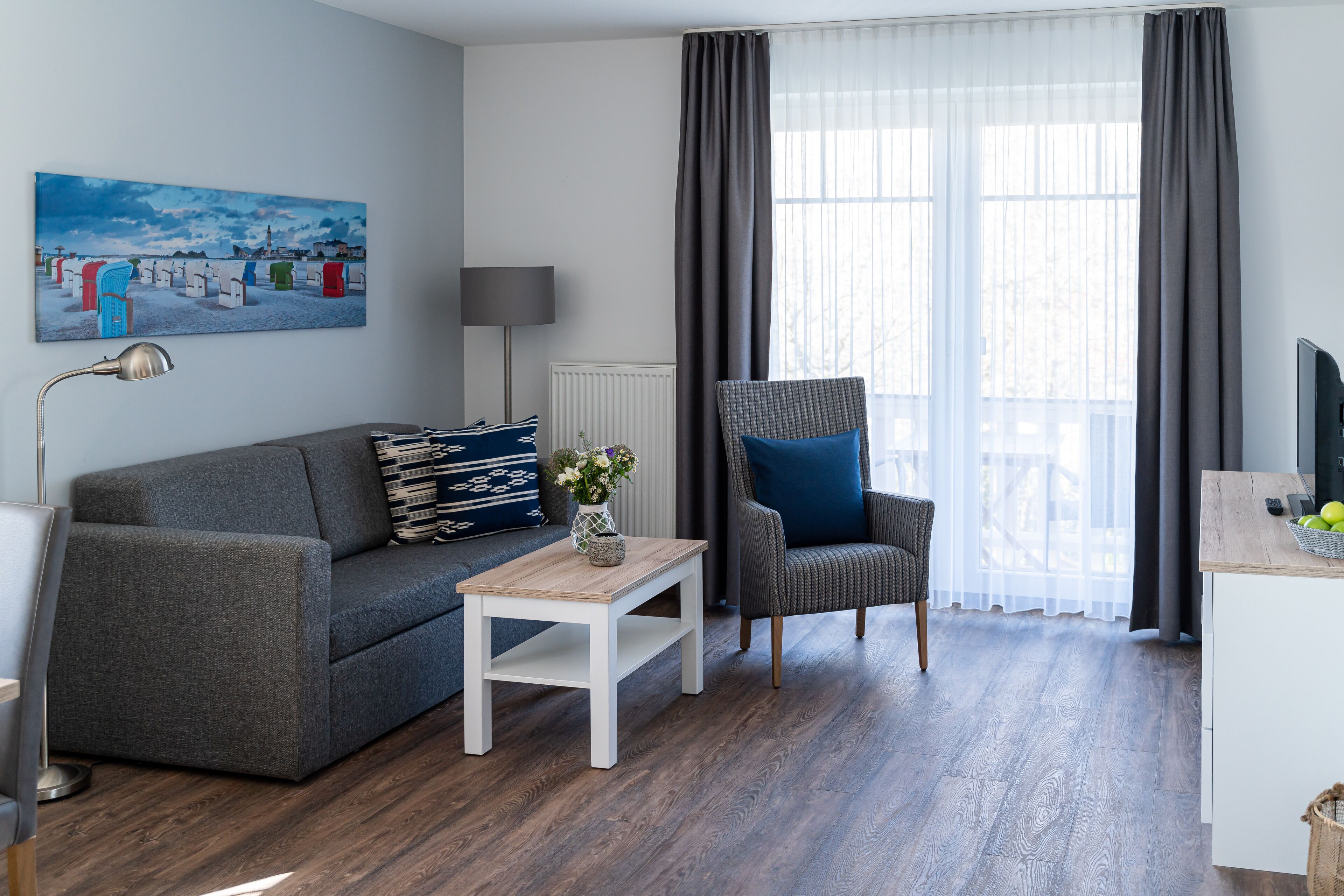 A modern living room with a grey sofa, a small white table with a vase, a grey armchair, and a painting on the wall.