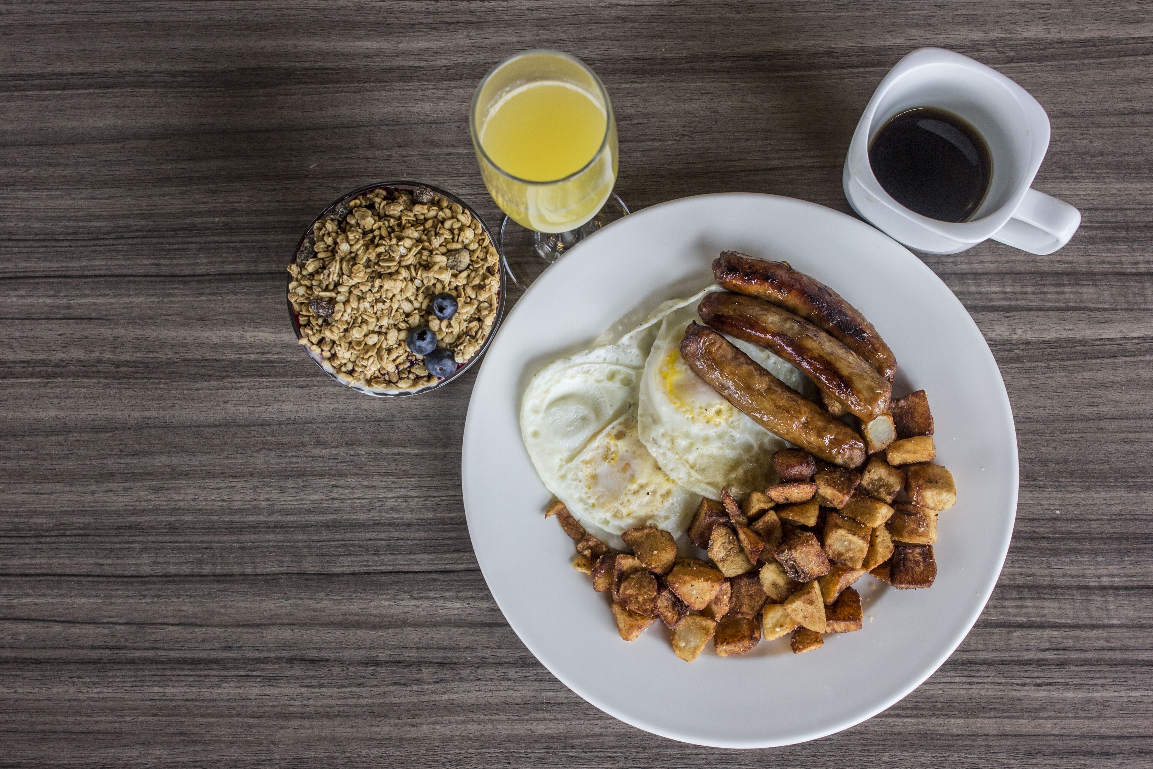 Un plato de desayuno con huevos, salchichas y papas, un tazón de granola con arándanos, un vaso de jugo de naranja y una taza de café sobre una mesa de madera.
